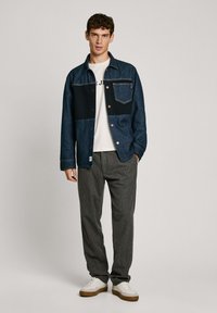 Giubbotto di jeans con toppe in diverse tonalità di blu, t-shirt bianca sotto, pantaloni a righe grigie e sneakers bianche.