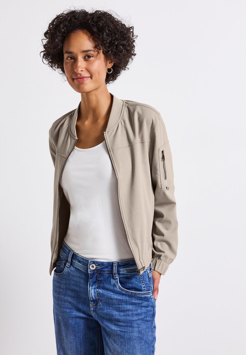 Jonge vrouw met krullend haar glimlacht, draagt een lichtgewicht beige bomberjack over een wit shirt en blauwe spijkerbroek, voor een effen achtergrond.