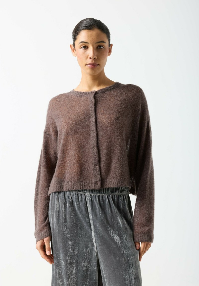 Junge Frau trägt eine braune, flauschige Strickjacke mit Knöpfen und lockere, graue Samthosen, steht vor einem einfarbigen hellen Hintergrund.