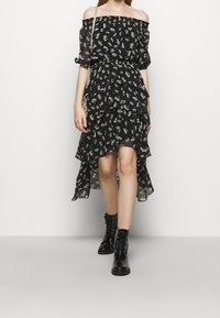 Schwarzes, blumiges Off-Shoulder-Kleid mit rüschenartigen Lagen, transparentem Material und knielanger Schnittführung, kombiniert mit schwarzen Schnürstiefeln.