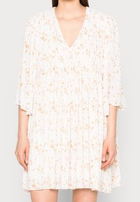 Femme portant une robe plissée blanche avec des motifs floraux beiges et des manches trois-quarts, se tenant devant un fond uni.