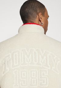 Veste en polaire crème avec un col haut, ornée d'une inscription en relief blanche "TOMMY 1985" au dos et d'une doublure intérieure rouge contrastante.