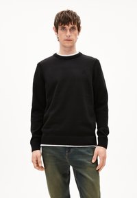 Schwarzer Pullover aus weichem Strickmaterial, mit Rundhalsausschnitt und geripptem Saum, versehen mit einem dezenten Logo-Detail auf der Brust. Darunter wird ein weißes Hemd getragen.