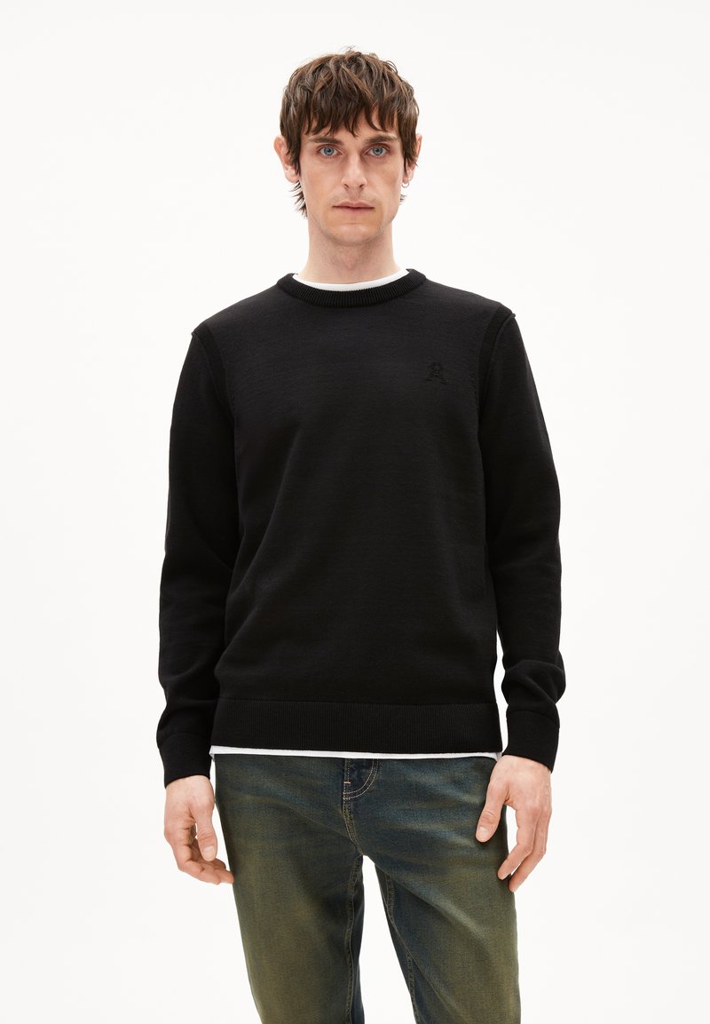 Schwarzer Pullover aus weichem Strickmaterial, mit Rundhalsausschnitt und geripptem Saum, versehen mit einem dezenten Logo-Detail auf der Brust. Darunter wird ein weißes Hemd getragen.