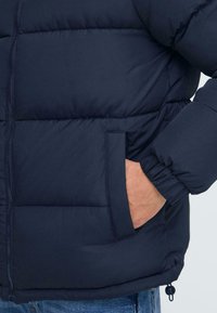 Marineblå polstret jakke med lynlåslukning, elastiske manchetter og sidelommer. Glat tekstur med kviltet design for isolering.