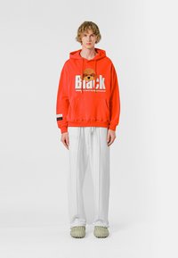 Sudadera roja con letras gráficas y un diseño de personaje, combinada con pantalones anchos blancos y zapatos de lona ligeros verde claro.