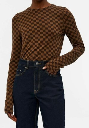 Osoba w brązowej i ciemnobrązowej koszulce w kratę z długim rękawem, włożonej do wysokich, ciemnoniebieskich dżinsów z przodu na guziki i kieszeniami.