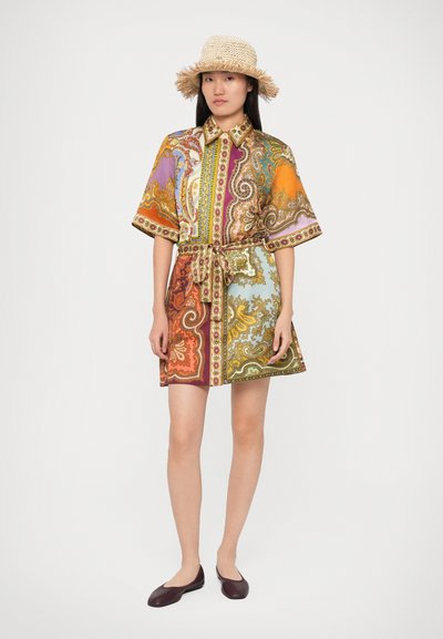 Femeie îmbrăcată într-o rochie cămașă beltată cu model paisley multicolor, pălărie din paie cu franjuri și pantofi negri, stând pe un fundal alb uniform.
