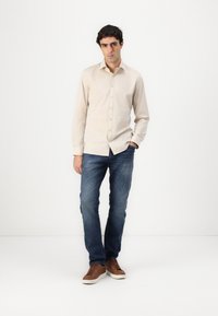 Cămașă bej cu butoni, cu guler, asortată cu blugi din denim albastru și adidași maro cu șireturi. Design simplu, formă ajustată, textură netedă.