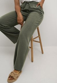 Green wide-leg trousers made of soft fabric, featuring an elastic waistband and side pockets. Worn with textured beige slide sandals.
