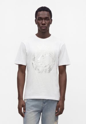 Junger Mann mit kurzen Haaren, der ein weißes T-Shirt mit Grafik und hellblaue, abgenutzte Jeans trägt, steht vor einem schlichten weißen Hintergrund.