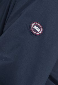 Navy fabric jacket featuring a round patch with "Jack & Jones" logo in white and red. Smooth texture, simple design, suitable for casual wear.