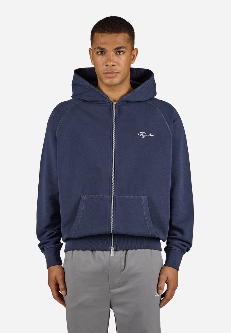 Man in een marineblauwe hoodie met rits en een capuchon, met voorzakken, gecombineerd met lichtgrijze broek tegen een effen achtergrond.