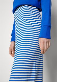 Jupe à rayures horizontales bleues et blanches portée avec un haut bleu à manches longues, montrant un gros plan du bas du torse et des mains.