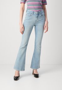 Jeans de mezclilla de corte acampanado en azul claro con un acabado desgastado, que presentan cinco bolsillos y una cinturilla con pasadores para el cinturón. Llevados con tacones de plataforma negros.