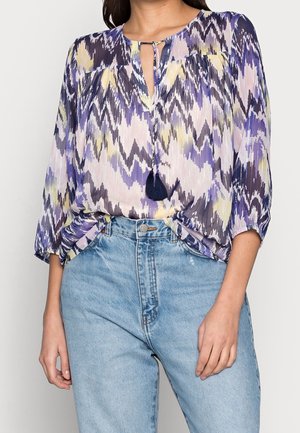 Blusa fluida nei toni di viola e giallo con motivi a zigzag. Presenta un colletto a fessura e una nappina decorativa. Tessuto leggero.