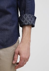 Bras avec manche remontée de chemise en denim bleu foncé montrant le tissu intérieur à motifs, reposant à côté d’un pantalon beige sur un fond uni.