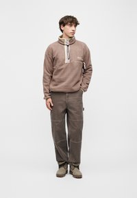 Νέος άνδρας που φοράει καφέ φλις πουλόβερ, φαρδιά καφέ παντελόνια και μπεζ μποτάκια, στέκεται με το ένα χέρι στην τσέπη μπροστά σε μια απλή φόντο.