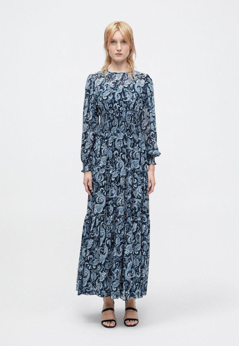 Μπλε μάξι φόρεμα με μοτίβο πεϊσλί, μακριά μανίκια, μπούστο με σφηκοφωλιά, φούστα με στρώσεις και μαύρα σανδάλια με λουριά. Απαλό, ρέον ύφασμα.