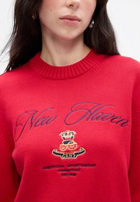 Rød strikket sweater med ribbet halsudskæring, broderet "New Haven" i blå og et våbenskjold med "GANT" samt tekst i guld og marineblå.