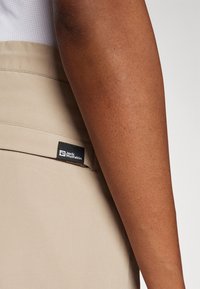Un tissu beige au toucher lisse présente une étiquette de marque noire, positionnée sur le côté gauche près d'une poche, avec un bras partiellement visible dans le cadre.