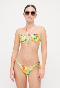 Floral bikini-setti vihreänä oranssilla ja keltaisilla kuvioilla. Olkaimeton yläosa, jossa on nauhakiinnitys, ja sivunauhoilla varustetut alaosat. Tasainen, joustava kangas.