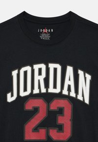 Čierne bavlnené tričko s okrúhlym výstrihom, na ktorom sa nachádza výrazný biely nápis "JORDAN" a červené číslo "23". Nápis má vrstvené prešívané okraje pre textúru.