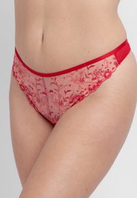 Εσώρουχο στυλ μπικίνι από κόκκινη δαντέλα με κεντήματα λουλουδιών, φορεμένο στους γοφούς και το κάτω μέρος της κοιλιάς μιας γυναίκας, σε ουδέτερο φόντο.