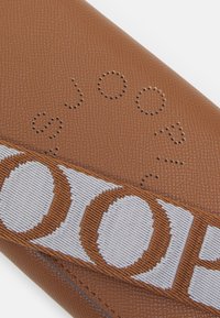 Brązowa, teksturowana skórzana torba z okrągłymi perforacjami oraz kontrastującym paskiem z napisem "JOOP!" w kolorze białym i brązowym.