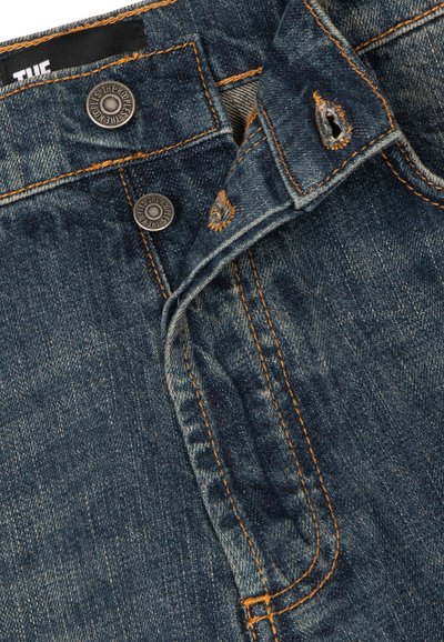 Denim jeans with two metallic buttons labeled "THE KOOPLES" and visible orange stitching on dark blue fabric.