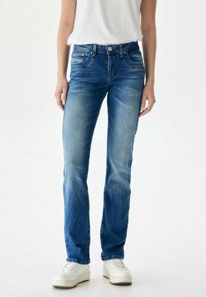 Vrouw met verweerde blauwe rechte jeans, witte sneakers en een wit mouwloos topje in haar broek gestopt, staand tegen een effen witte achtergrond.