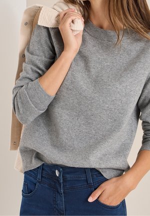 Woman wearing a grey knit sweater with ribbed cuffs and hem, dark blue jeans, and holding a beige corduroy jacket over her shoulder.