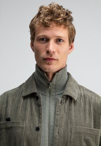 Olivenfarbene Cordjacke mit hohem Kragen, Reißverschluss vorne, gerippter Textur und aufgesetzter Tasche auf der linken Brust.