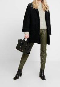 Svart överdimensionerad kappa, ljusgrå stickad tröja, gröna läderbyxor och svarta klackstövlar. Håller en svart handväska med guldlogotyp.