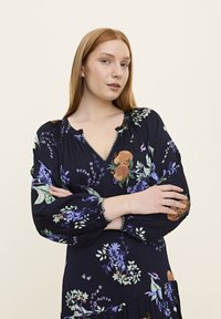 Blumen-Maxikleid mit navy Hintergrund, ausgestattet mit Puffärmeln, einem gewellten Ausschnitt und bunten botanischen Mustern in Grün und Lila.