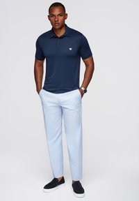Uomo in piedi con le mani nelle tasche di pantaloni azzurri chiari, indossa una maglietta polo blu navy, scarpe slip-on nere e un orologio d'argento, su sfondo bianco.