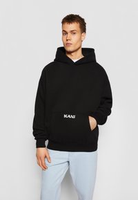 Black hoodie made of soft fabric, featuring a front pocket and embroidered logo. Paired with light blue jeans, showcasing a relaxed fit.