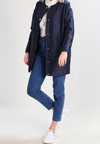 Osoba v námořnickém prošívaném kabátu, bílém tričku, modrých džínách a bílých teniskách, stojící s jednou nohou ohnutou proti světlému pozadí.