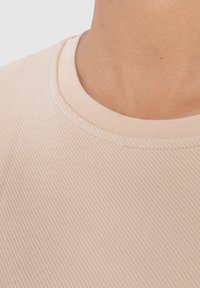 Vaaleanbeige tekstuuri kankaan lähikuva, jossa on röyhelökuvio, pyöreä pääntie ja sileät saumat. Pehmeä, muotoon puristettu malli.