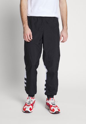 Man wearing black track pants with white stripes on the sides and white, red, and black Adidas sneakers, standing against a plain background.