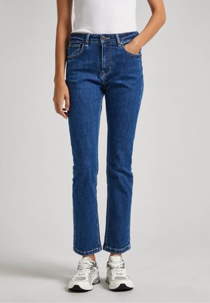 Femme portant un jean droit bleu avec des poches avant, associé à des baskets blanches et un haut blanc sans manches, debout devant un fond uni.