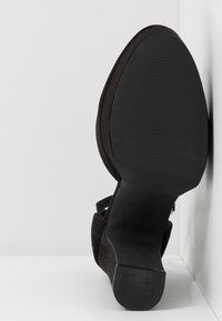 Chaussure noire à talon reposant sur le dos, montrant la semelle lisse et texturée ainsi que le talon carré sur fond blanc dans un coin.