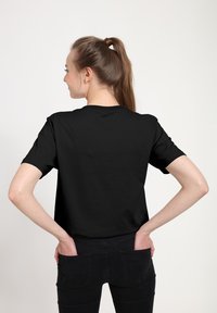 Černé bavlněné tričko s krátkými rukávy, jednoduchým kulatým výstřihem a volným střihem, doplněné černými džíny a jednoduchým pozadím.