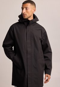 Parka negra impermeable con alto collar, cierre automático en la parte delantera, grandes bolsillos y una textura suave, adecuada para diversas condiciones climáticas.
