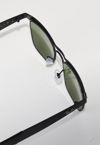 Ochelari de soare cu rame negre din metal, lentile verzi și pernițe pentru nas transparente. Au un design elegant și o marcă subtilă pe tâmplă.