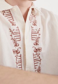 Krémová košile na knoflíky s propracovanou výšivkou v rustikálních barvách květin a pruhů podél předního zapínání a límce. Hladká textura tkaniny.