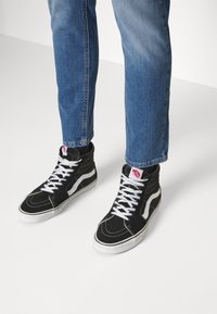 Svarta hög-topp sneakers i canvas, med vita snören, kontrasterande vit sidremsa och en röd logotagg på tungan.