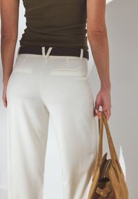 Balti platās bikses ar diviem aizmugurējiem kabatām, kombinētas ar tumšu pītā jostu. Labajā rokā tur tan krāsas zamšādas rokassomu.