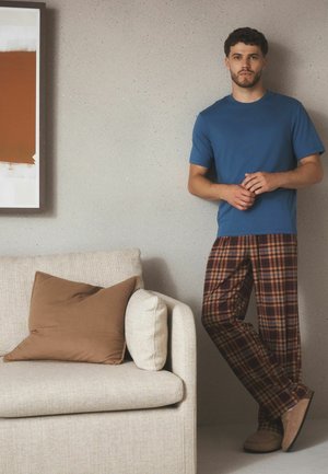 Man in blauw overhemd en geruite broek leunt tegen een lichtgrijze muur naast een beige bank met bruine en crèmekleurige kussens.