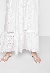 Robe en coton blanc avec broderie anglaise, ourlet au genou et volant. Associée à des sandales blanches à lanières laissant les orteils visibles.